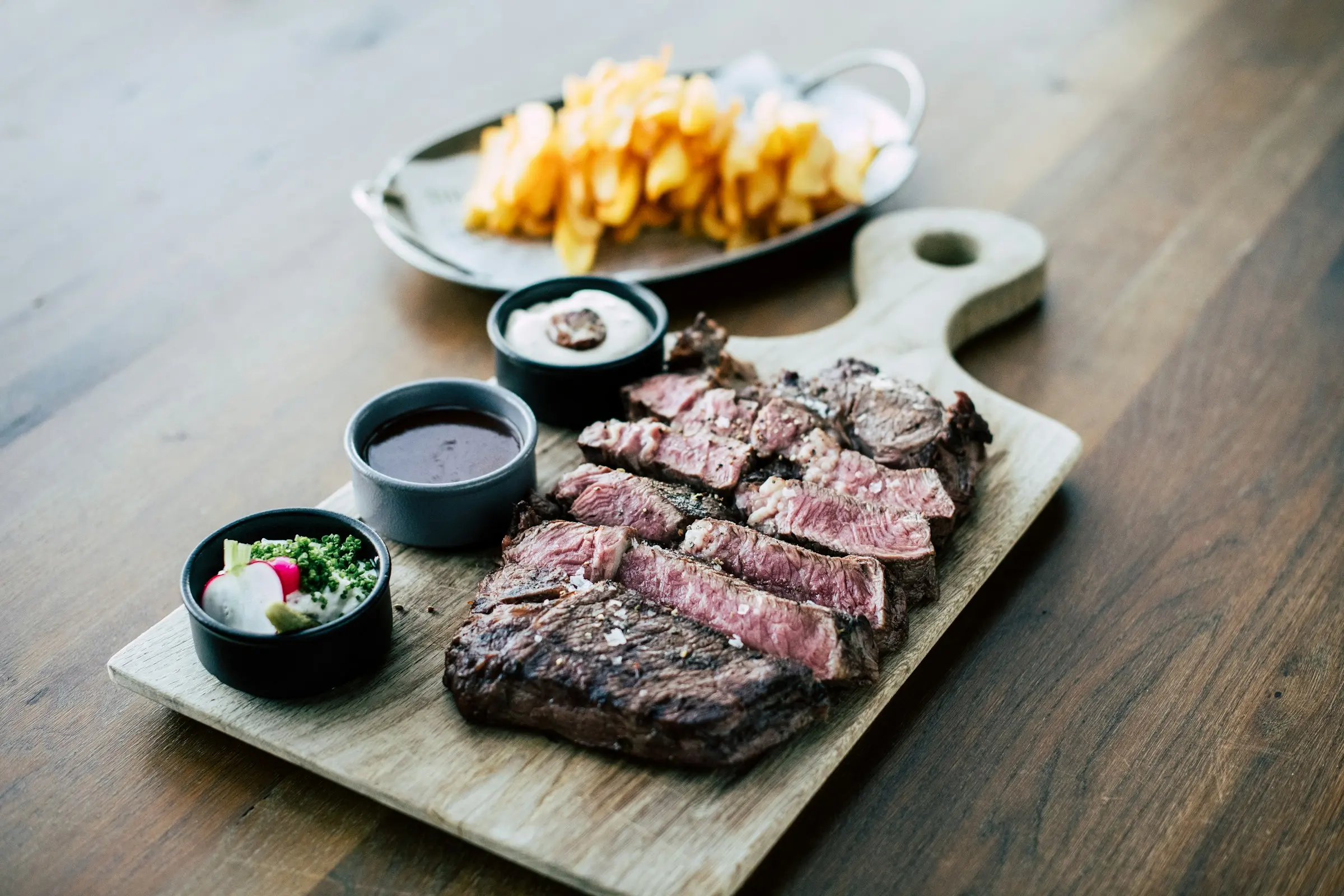 Sliced grilled steak on a board with dipping sauces and fries